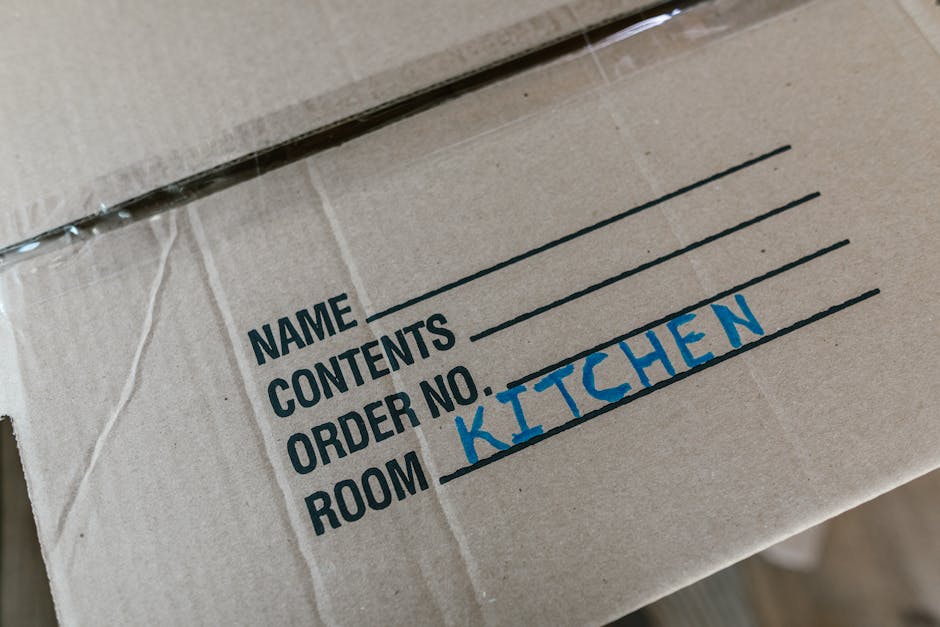 A close-up photograph of a cardboard moving box with printed labels indicating fields for 'Name', 'Contents', 'Order No.', and 'Room', which has been handwritten in blue ink as 'Kitchen'. The box is p