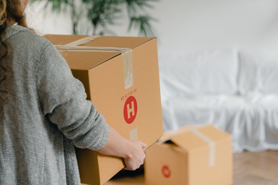 A close-up photograph of a cardboard moving box with printed labels indicating fields for 'Name', 'Contents', 'Order No.', and 'Room', which has been handwritten in blue ink as 'Kitchen'. The box is p